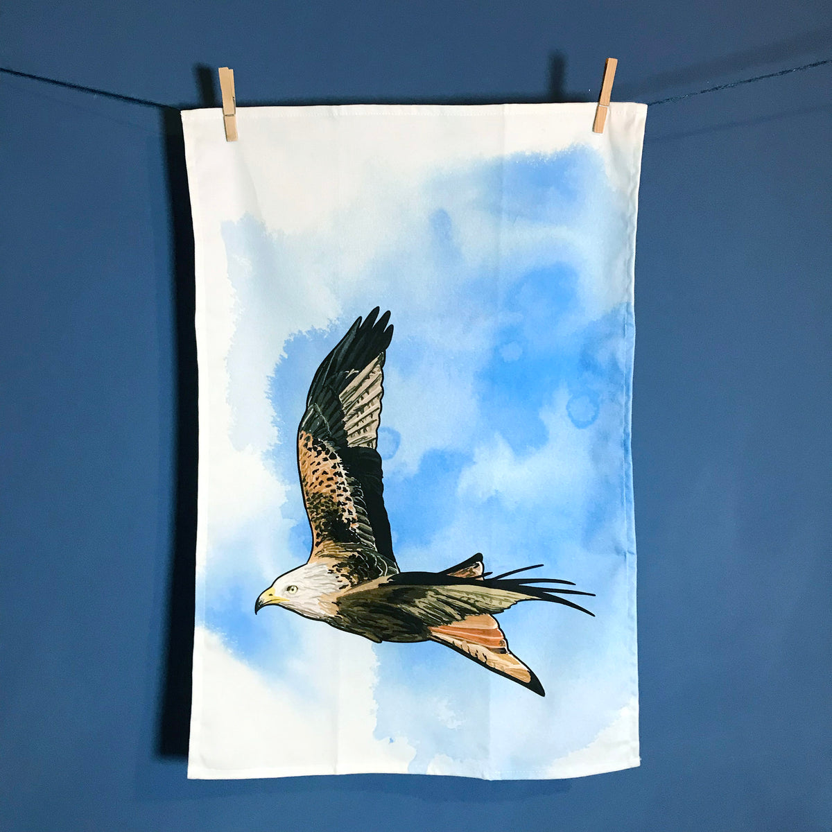 A  photo of tea towel hanging on a washing line against a dark blue wall. The tea towel shows an illustration of a flying red kite against an inky bright blue wash white background.