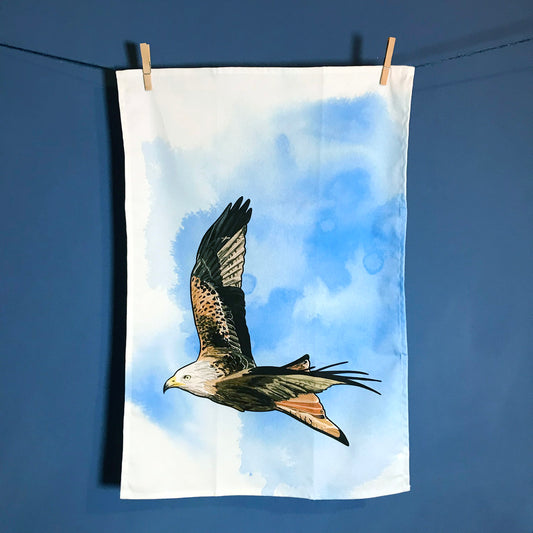 A  photo of tea towel hanging on a washing line against a dark blue wall. The tea towel shows an illustration of a flying red kite against an inky bright blue wash white background.