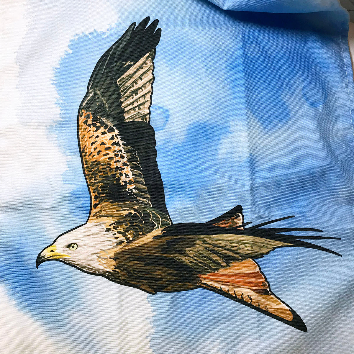 A close up photo of a tea towel showing an illustration of a flying red kite against an inky wash bright blue and white background.