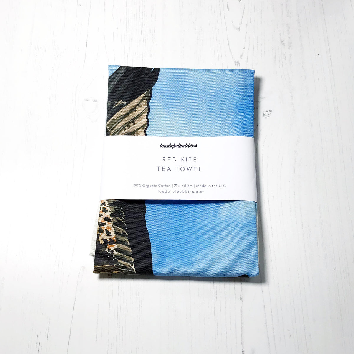 A folded bird themed illustration tea towel by loadofolbobbins. It shows a flying red kite against an inky wash bright blue and white background.