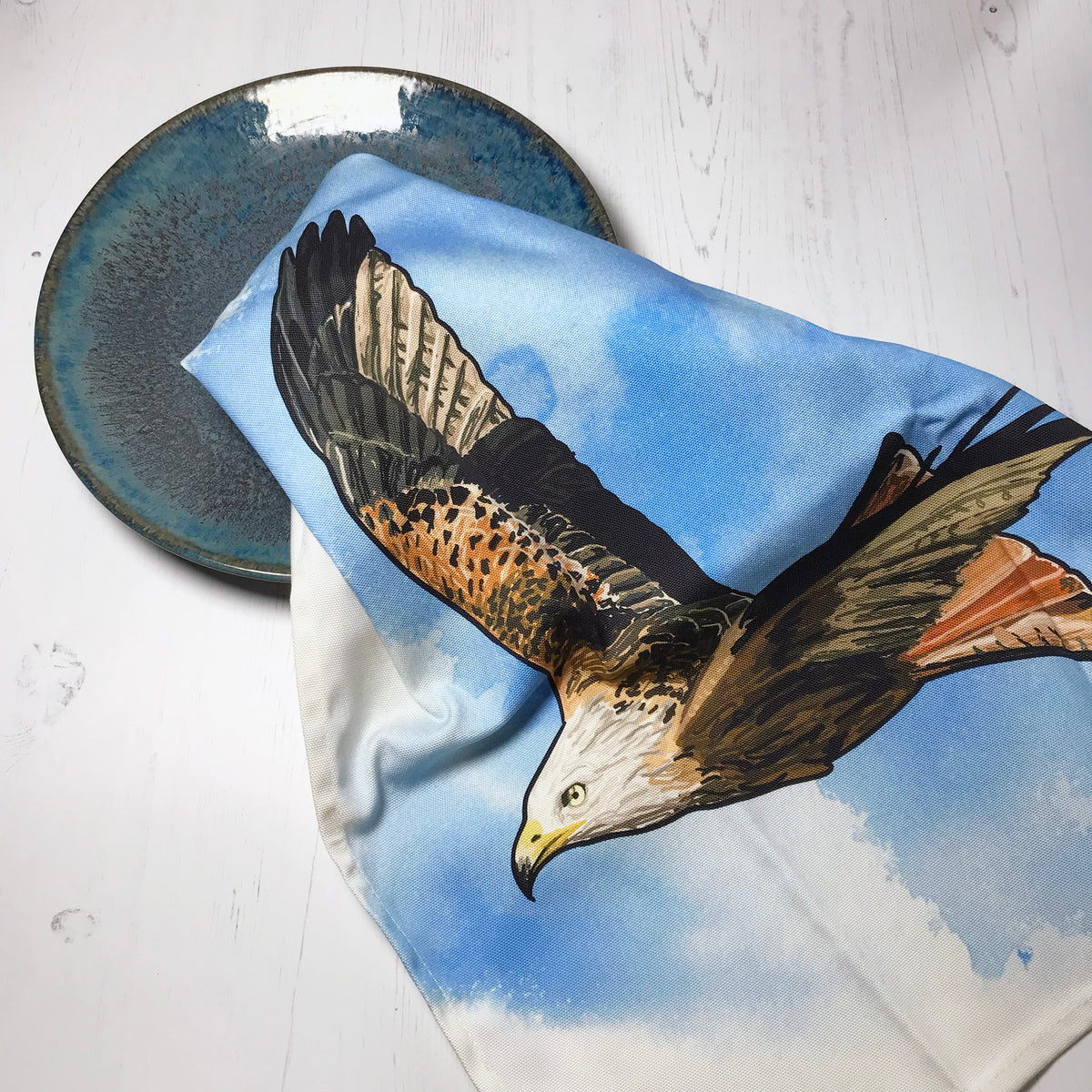 A  photo of a loosely scrunched up tea towel looking like it is drying a ceramic glazed dark turquoise plate. The tea towel shows an illustration of a flying red kite,  against an inky wash bright blue and white background.