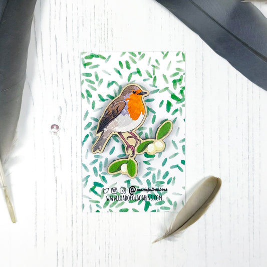 A laser cut wooden brooch showing an illustration by Loadofolbobbins of a robin bird perched on some mistletoe. The brooch is mounted on a backing card with a repeat abstract watercolour leaf pattern.