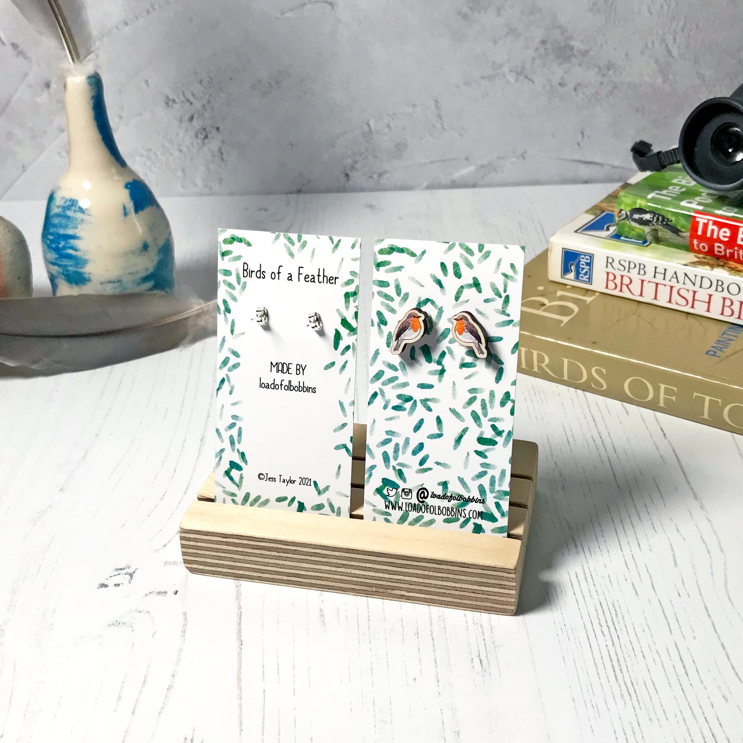 A pair of laser cut wooden earrings showing an illustration of a robin bird by loadofolbobbins. The earrings are mounted on a mini backing card with a repeat abstract watercolour leaf pattern. They are standing in a wooden stand and in the background you can see a pile of bird watching books and a pair of binoculars.