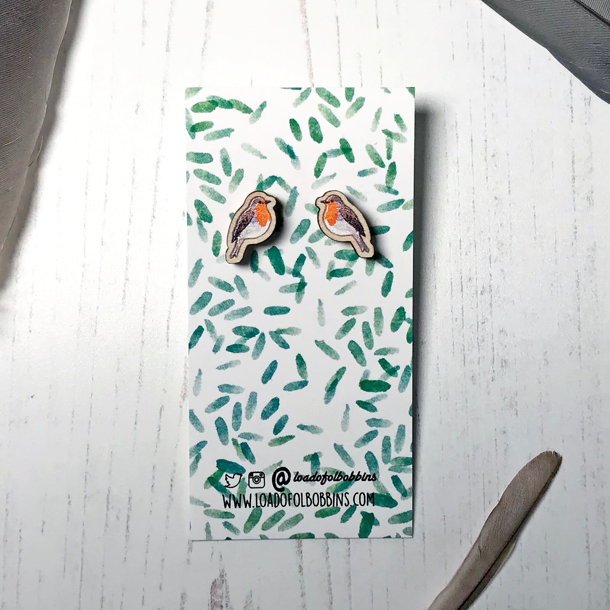 A pair of laser cut wooden earrings showing an illustration by Loadofolbobbins of a robin bird. The earrings are mounted on a mini backing card with a repeat abstract watercolour leaf pattern.
