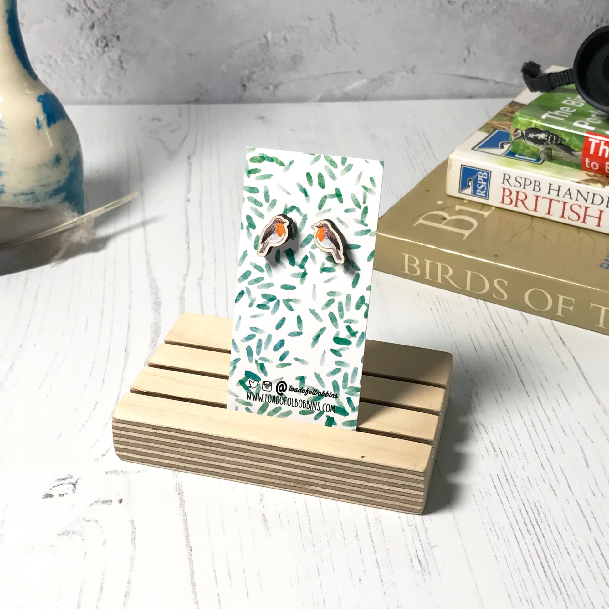 A pair of laser cut wooden earrings showing an illustration of a robin bird by loadofolbobbins. The earrings are mounted on a mini backing card with a repeat abstract watercolour leaf pattern. They are standing in a wooden stand and in the background you can see a pile of bird watching books and a pair of binoculars.