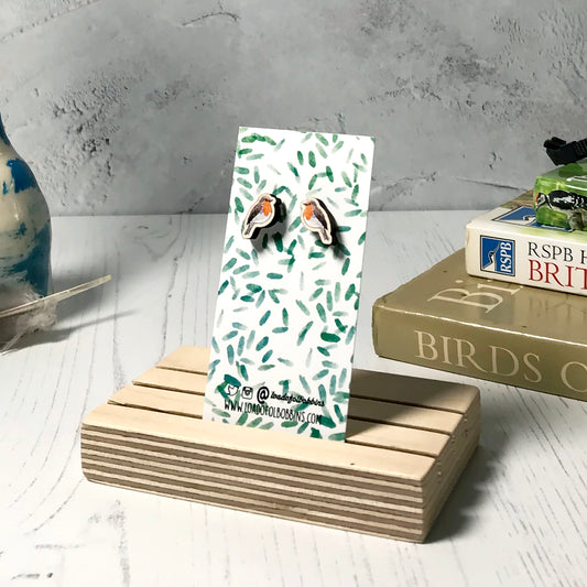 A pair of laser cut wooden earrings showing an illustration of a robin bird by loadofolbobbins. The earrings are mounted on a mini backing card with a repeat abstract watercolour leaf pattern. They are standing in a wooden stand and in the background you can see a pile of bird watching books and a pair of binoculars.