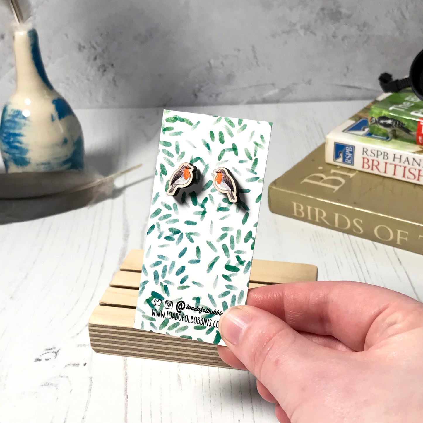 A pair of laser cut wooden earrings showing an illustration of a robin bird by loadofolbobbins. The earrings are mounted on a mini backing card with a repeat abstract watercolour leaf pattern. They are being held by a hand for scale, which shows that each stud earring is slightly smaller than a thumbnail.