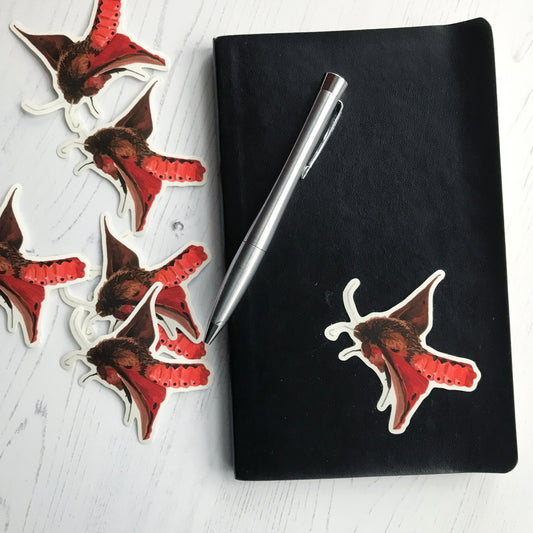 A ruby tiger moth sticker created from an original digital illustration by Loadofolbobbins. It has a rich brown very fluffy upper body and head with a vibrant red segmented lower abdomen with a black diamond pattern running down the centre, and a series of black dots along the side. It's wings have rich reddish brown tones with black spot markings. It's antennae are a light almost silver grey colour. The view of the moth is side on and looks like it is just taking off in flight.