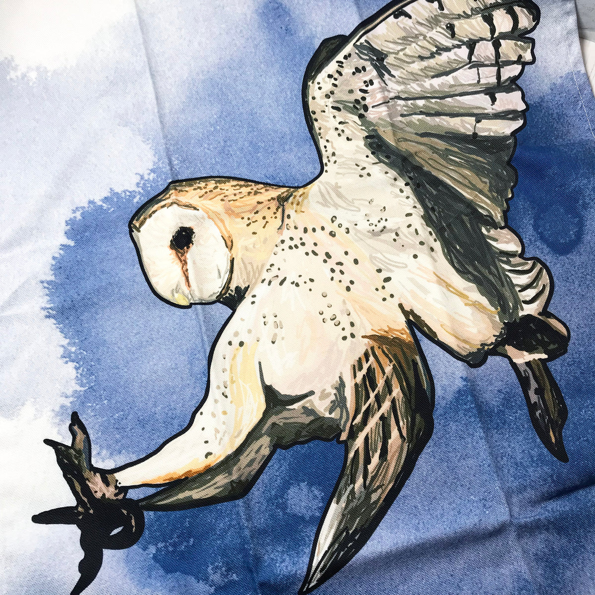 A close up photo of a tea towel showing an illustration of a swooping barn owl against an inky wash background.