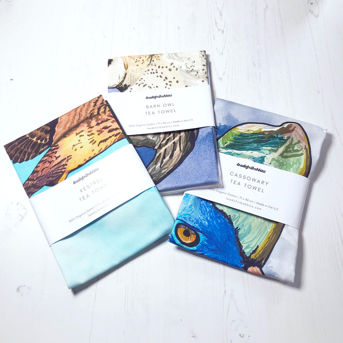 A collection of bird themed illustration tea towels by loadofolbobbins. Three folded tea towels one shows a hovering kestrel, a swooping barn owl and the head of a cassowary bird.