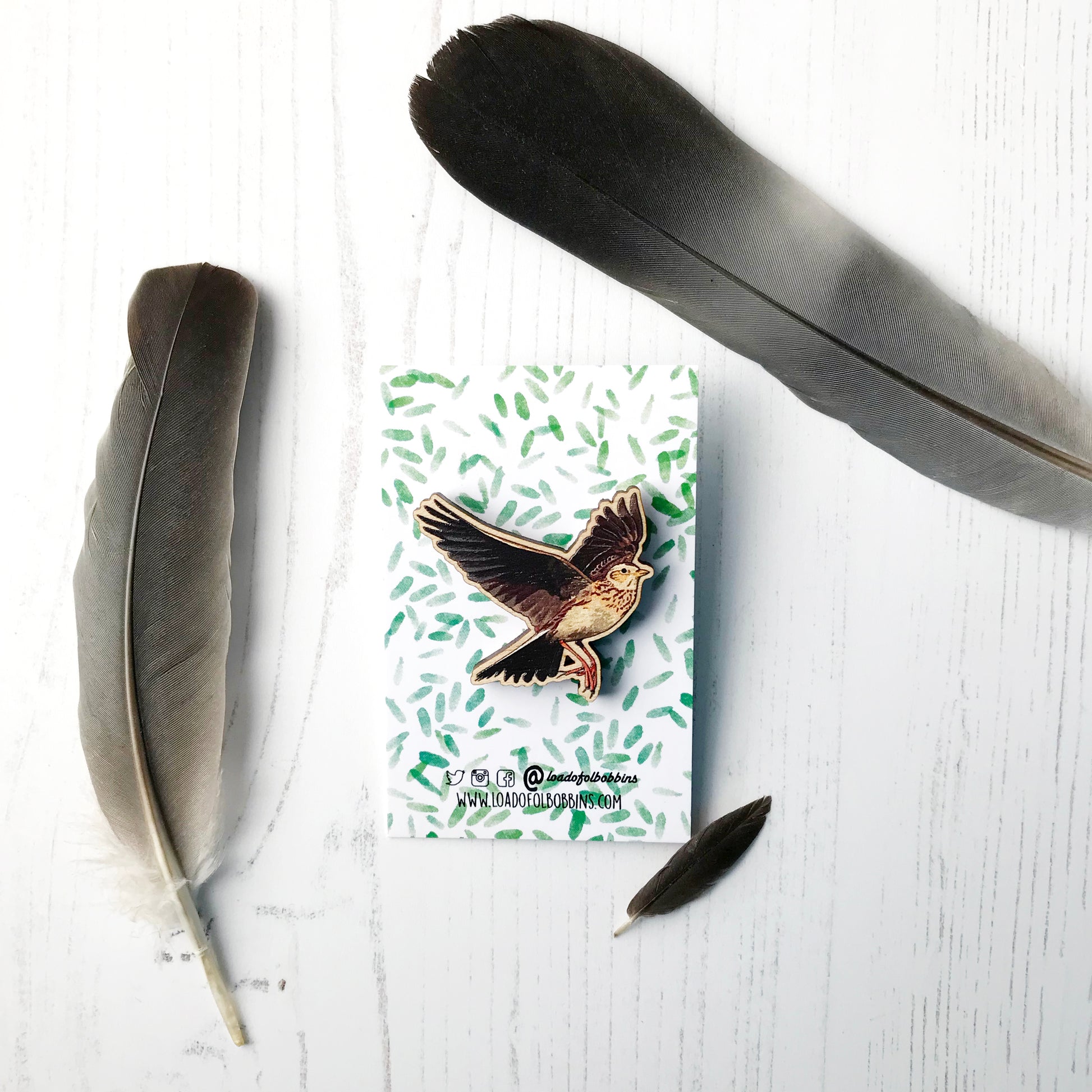 A laser cut wooden brooch showing an illustration by Loadofolbobbins of a flying skylark with wings spread out wide. The brooch is mounted on a backing card with a repeat abstract watercolour leaf pattern.