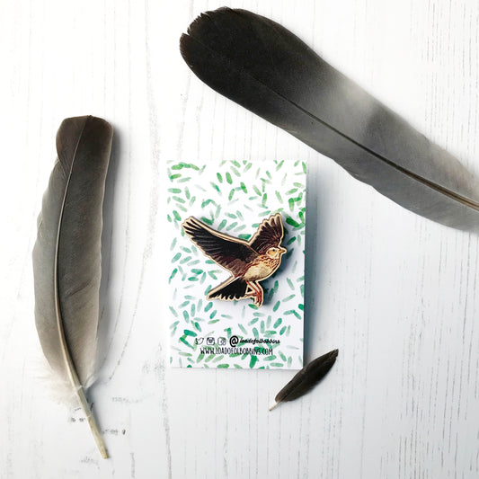 A laser cut wooden brooch showing an illustration by Loadofolbobbins of a flying skylark with wings spread out wide. The brooch is mounted on a backing card with a repeat abstract watercolour leaf pattern.
