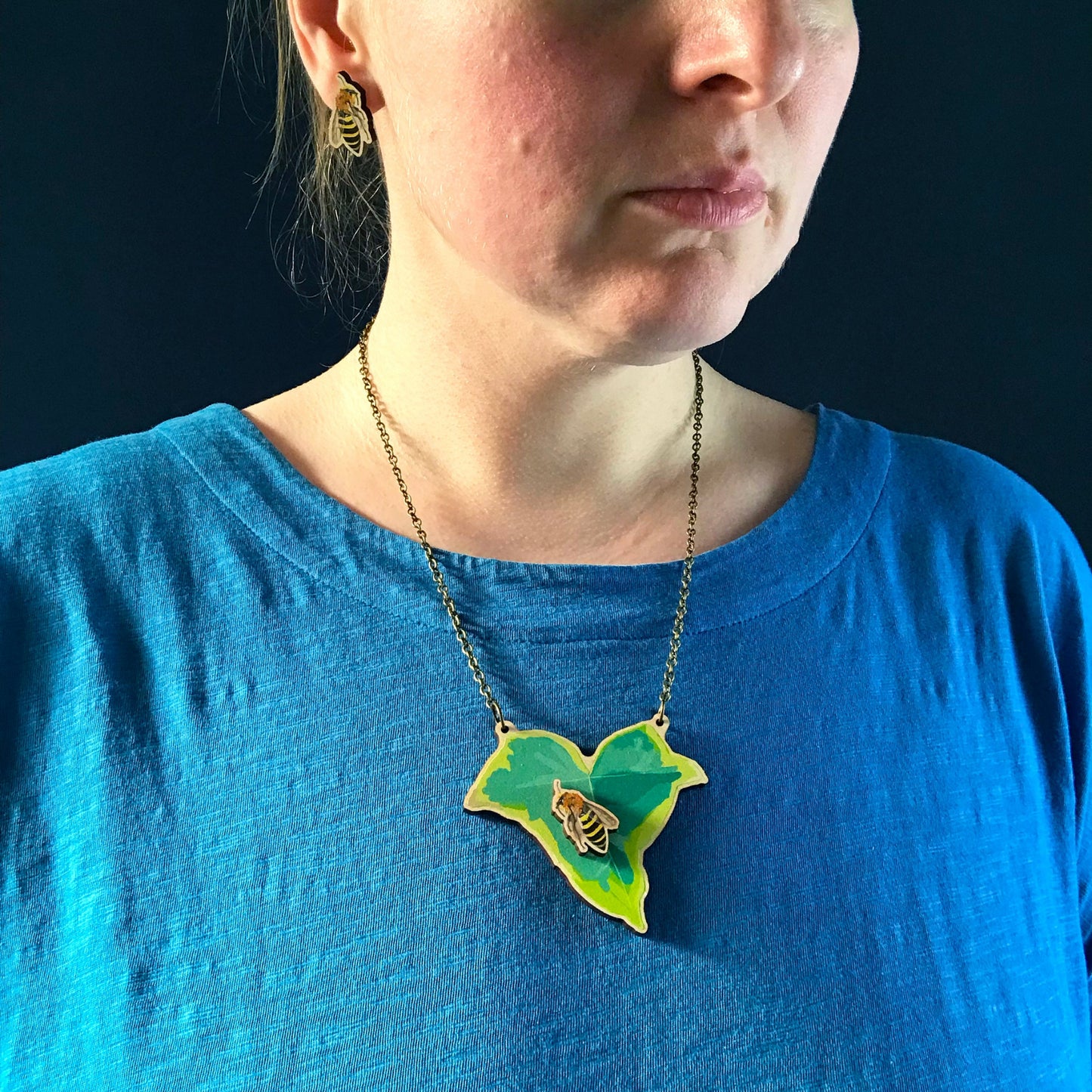 Small Ivy and Mining Bee  necklace by Loadofolbobbins. A laser cut wooden necklace that shows a small ivy leaf with an ivy mining bee on it. The necklace hangs on an antique bronze chain. It is being worn by me a white woman wearing a light blue top. You can just see the bottom of my face and neck.