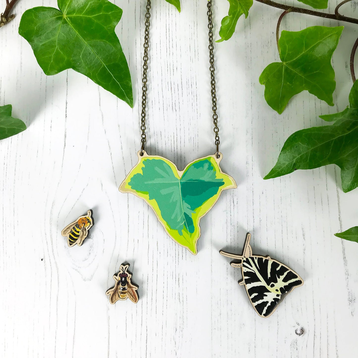 A laser cut wooden statement necklace showing an illustration of an variegated  green ivy leaf by loadofolbobbins. The wooden necklace is hanging from an antique bronze cable chain. It is surrounded by wooden charms also made from illustrations from left to right they show an ivy mining bee, golden hoverfly and a Jersey Tiger moth.