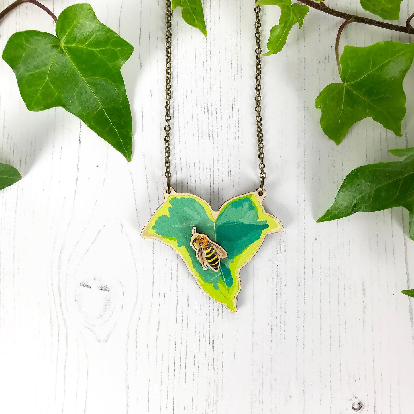 A laser cut wooden statement necklace showing an illustration of an variegated ivy leaf and an ivy mining bee by loadofolbobbins. The bee stands proud of the necklace in a 3D relief effect.  The wooden necklace is hanging from an antique bronze cable chain.