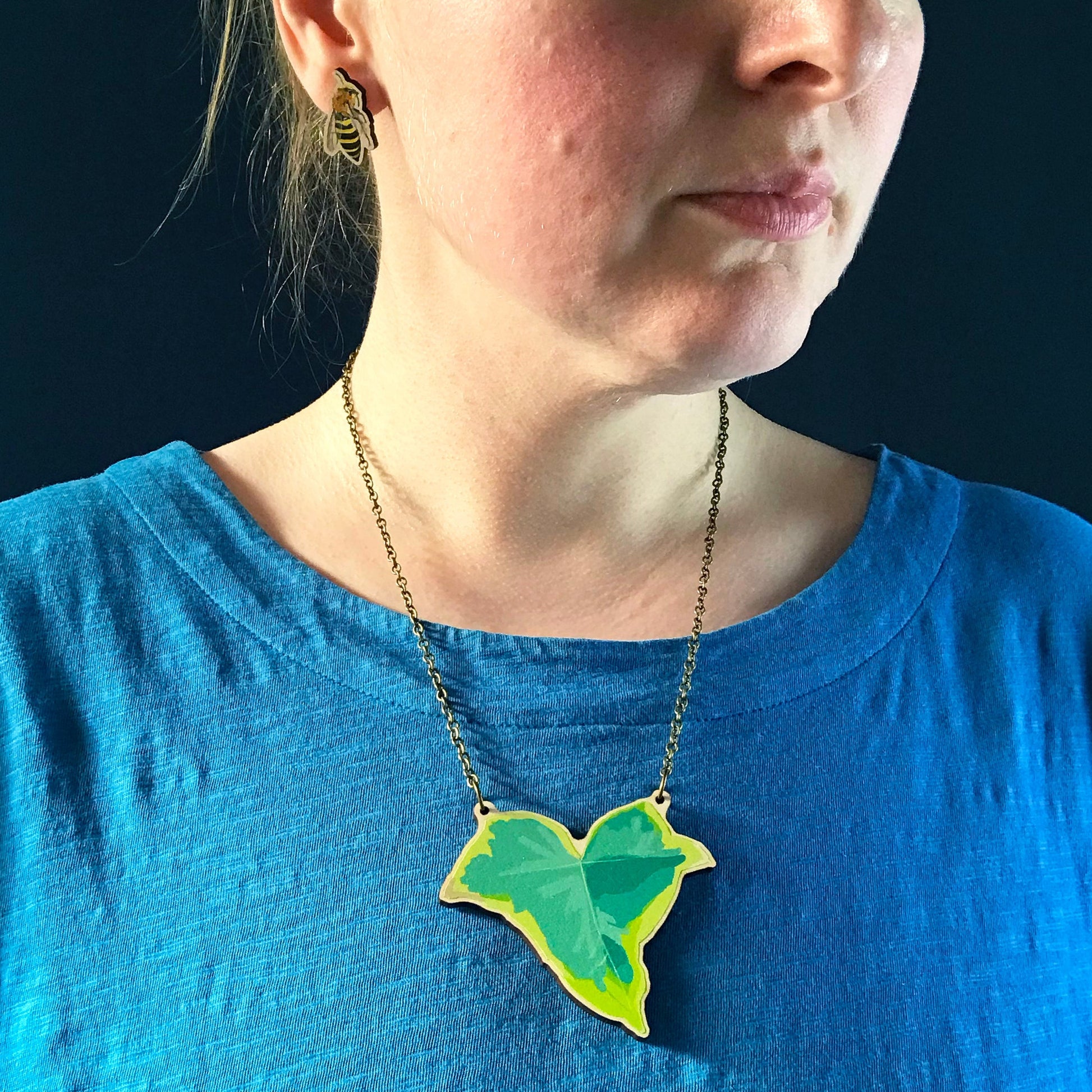 Small Ivy  necklace by Loadofolbobbins. A laser cut wooden necklace that shows a small variegated ivy leaf. The necklace hangs on an antique bronze chain. It is being worn by me a white woman wearing a light blue top. You can just see the bottom of my face and neck.