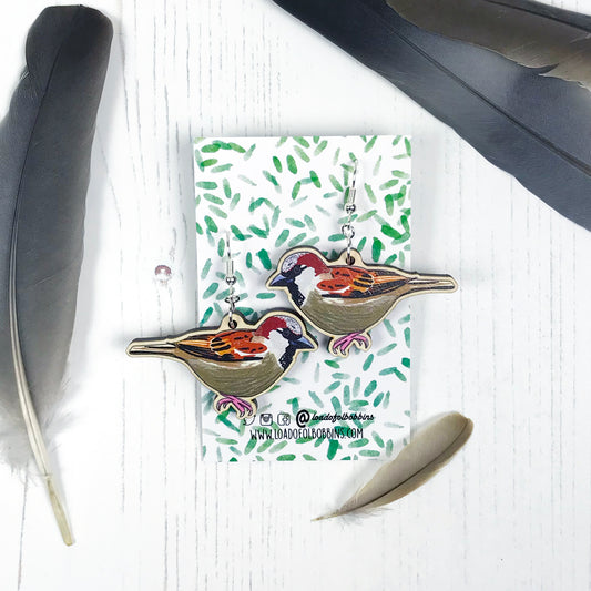 A pair of laser cut wooden dangle earrings showing an illustration by Loadofolbobbins of a male sparrow. The earrings are mounted on a backing card with a repeat abstract watercolour leaf pattern.