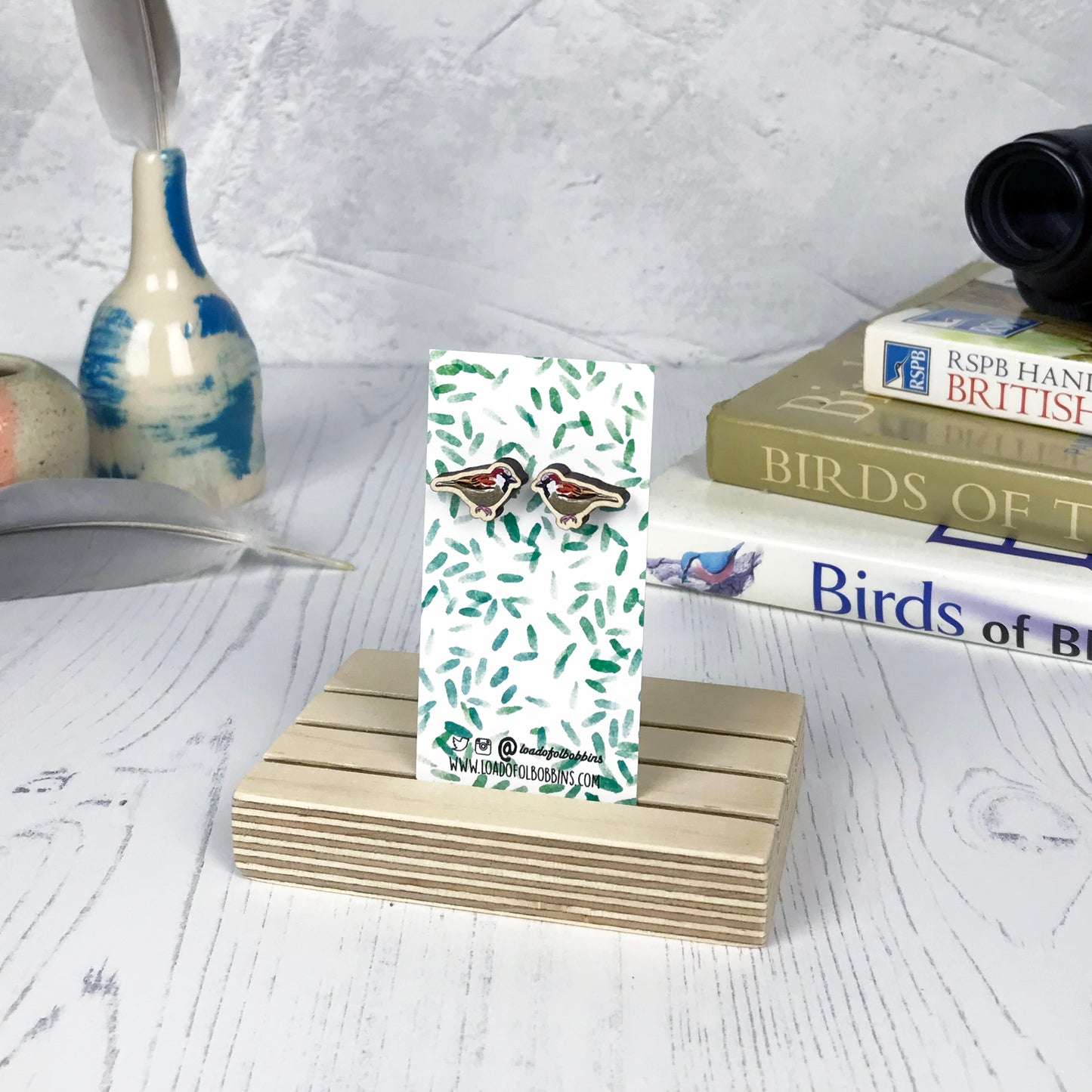 A pair of laser cut wooden earrings showing an illustration of a male sparrow bird by loadofolbobbins. The earrings are mounted on a mini backing card with a repeat abstract watercolour leaf pattern. They are standing in a wooden stand and in the background you can see a pile of bird watching books and a pair of binoculars.