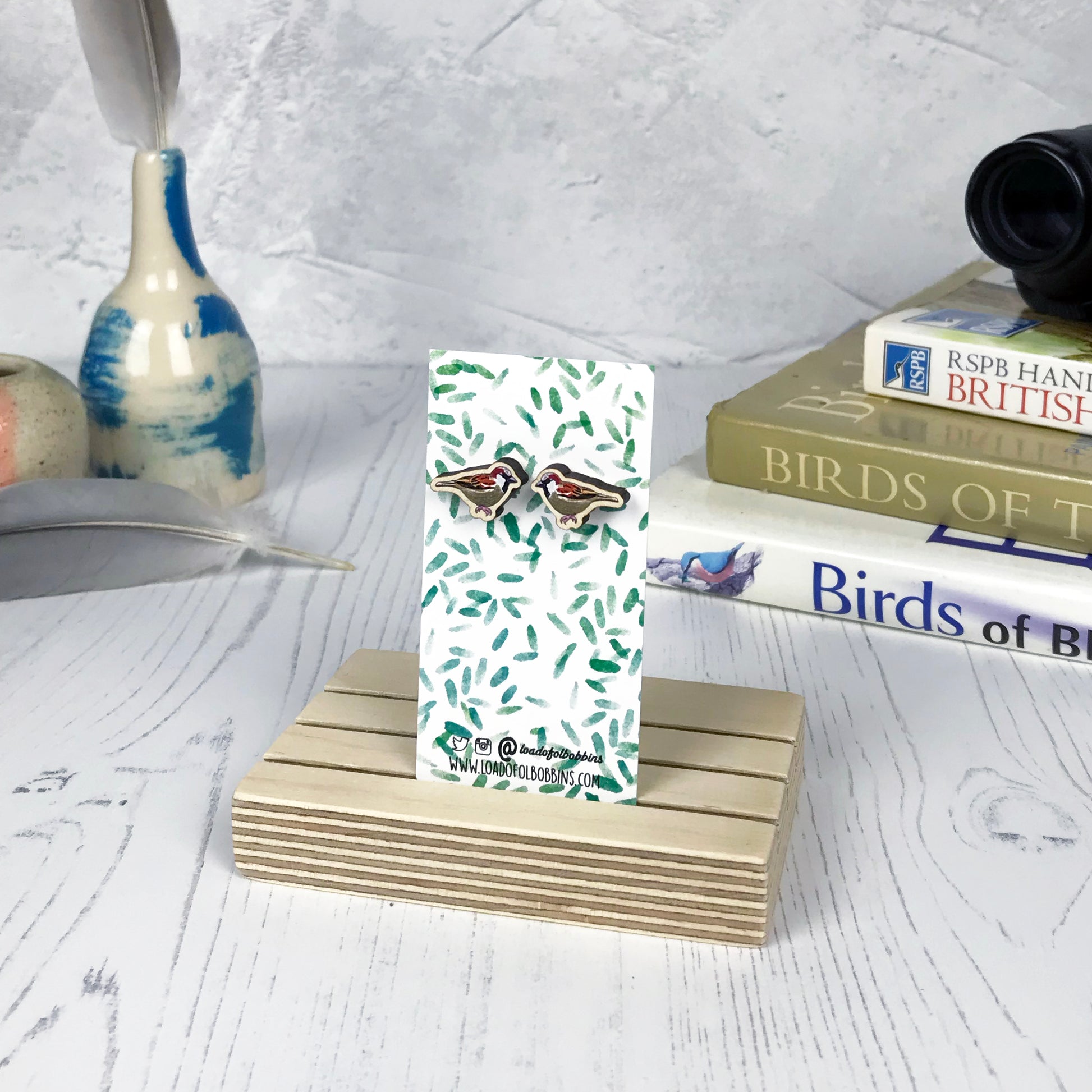 A pair of laser cut wooden earrings showing an illustration of a male sparrow bird by loadofolbobbins. The earrings are mounted on a mini backing card with a repeat abstract watercolour leaf pattern. They are standing in a wooden stand and in the background you can see a pile of bird watching books and a pair of binoculars.