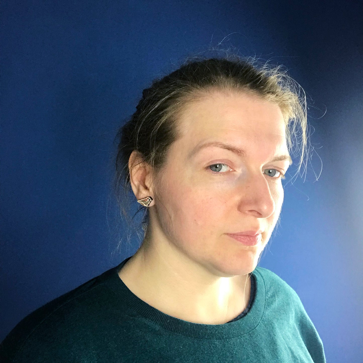 A neck and head close up photo of a white woman with blondish brown hair worn up and standing against a dark blue wall. Her face is slightly turned away from the camera so the main focus of the image is her ear which is wearing a small laser cut wooden stud earring of a male sparrow by Loadofolbobbins.