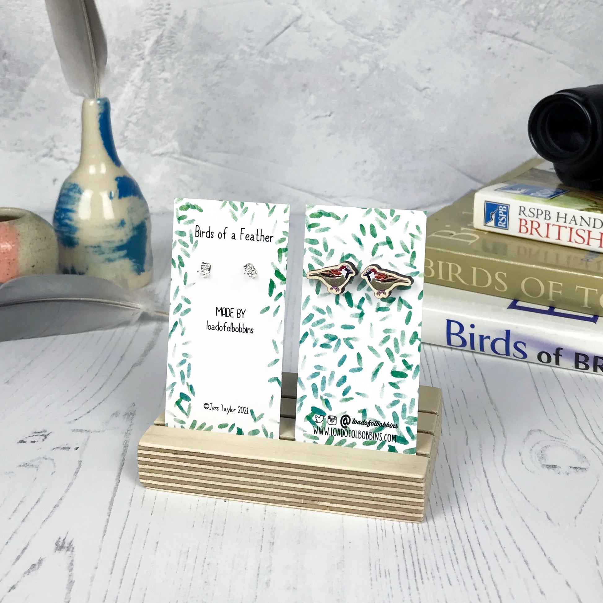 A pair of laser cut wooden earrings showing an illustration of a male sparrow bird by loadofolbobbins. The earrings are mounted on a mini backing card with a repeat abstract watercolour leaf pattern. They are standing in a wooden stand and in the background you can see a pile of bird watching books and a pair of binoculars.