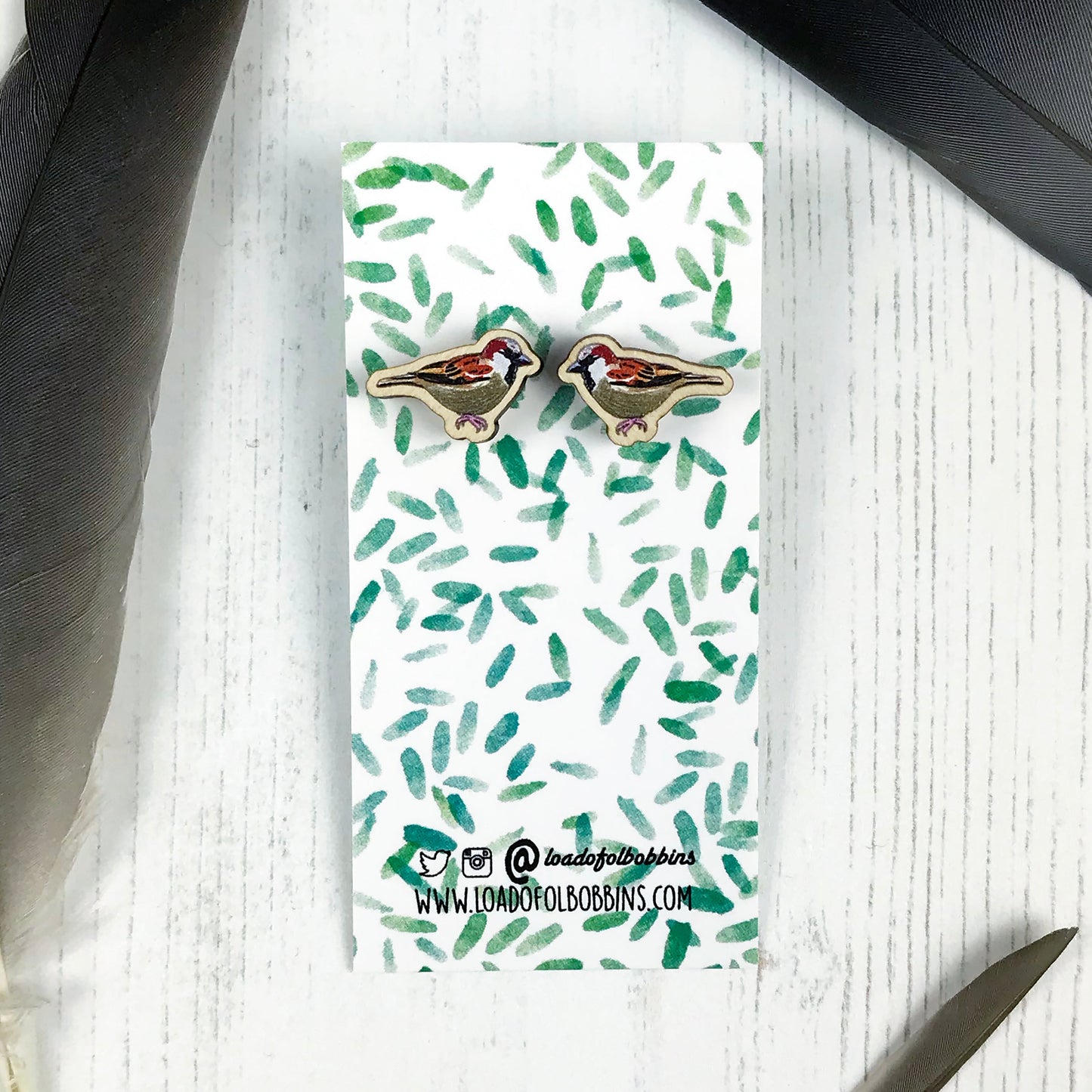 A pair of laser cut wooden earrings showing an illustration by Loadofolbobbins of a male sparrow bird. The earrings are mounted on a mini backing card with a repeat abstract watercolour leaf pattern.