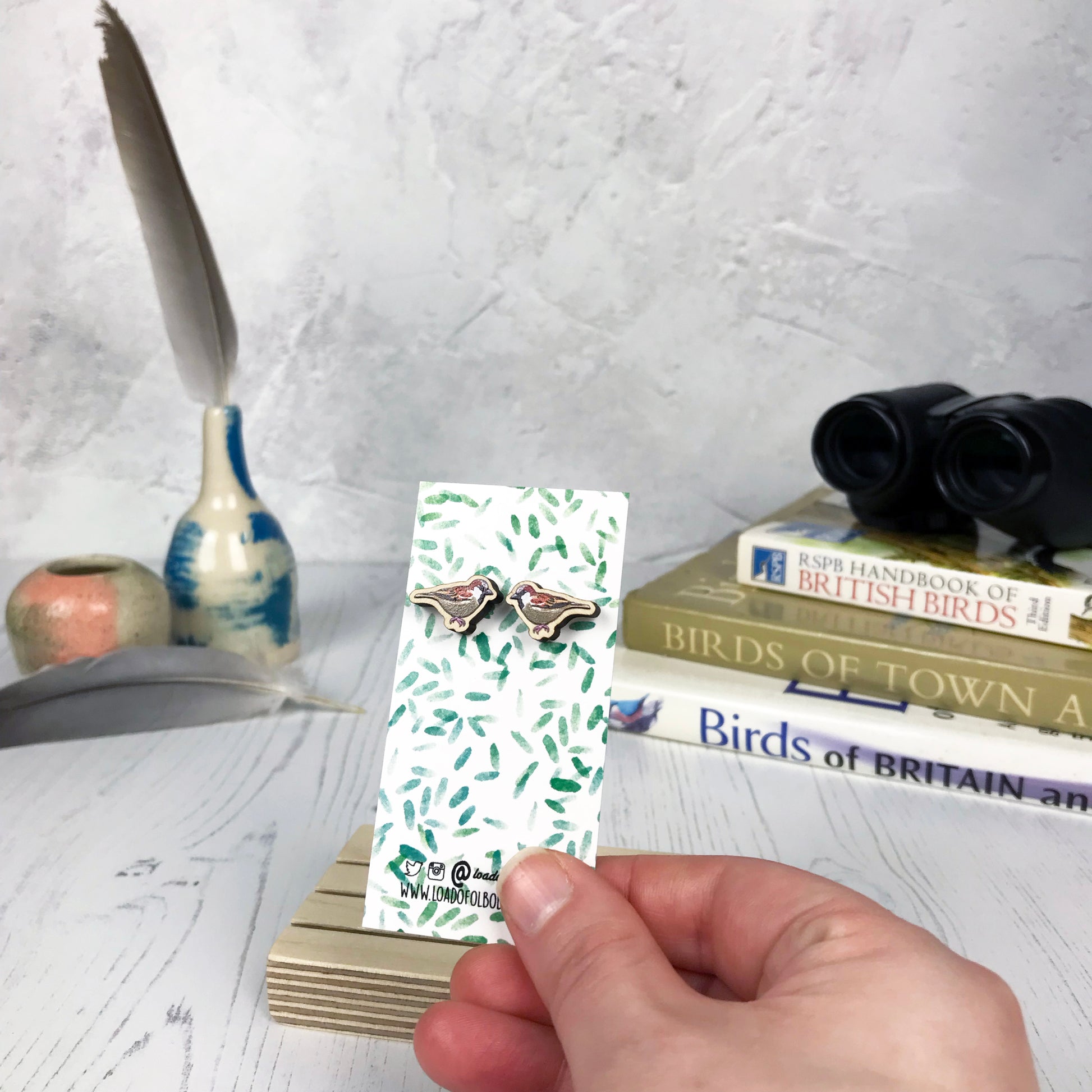 A pair of laser cut wooden earrings showing an illustration of a male sparrow bird by loadofolbobbins. The earrings are mounted on a mini backing card with a repeat abstract watercolour leaf pattern. They are being held by a hand for scale, which shows that each stud earring is slightly smaller than a thumbnail.
