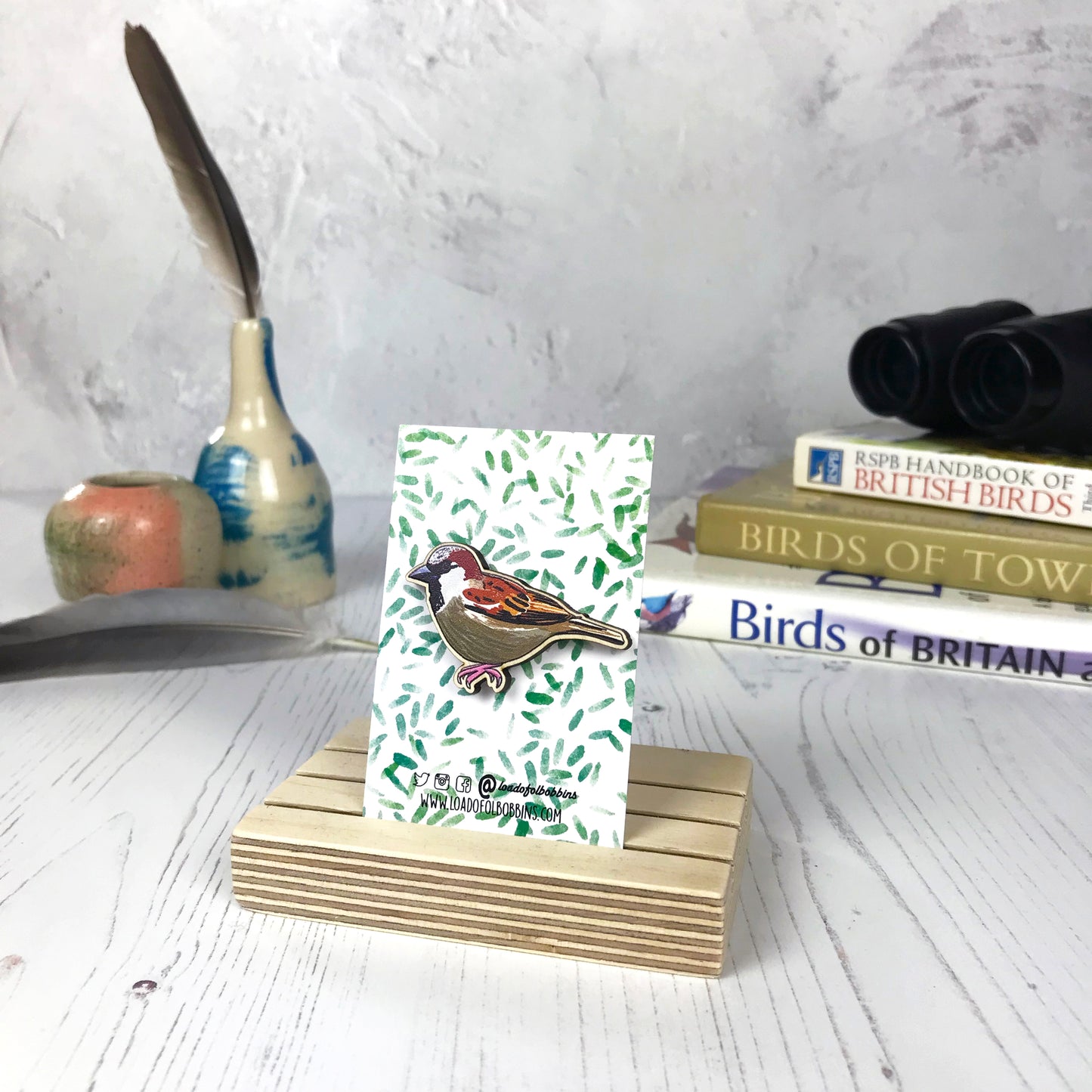 A laser cut wooden brooch showing an illustration of a male sparrow by loadofolbobbins. The brooch is mounted on a backing card with a repeat abstract watercolour leaf pattern. They are standing in a wooden stand and in the background you can see a pile of bird watching books and a pair of binoculars.