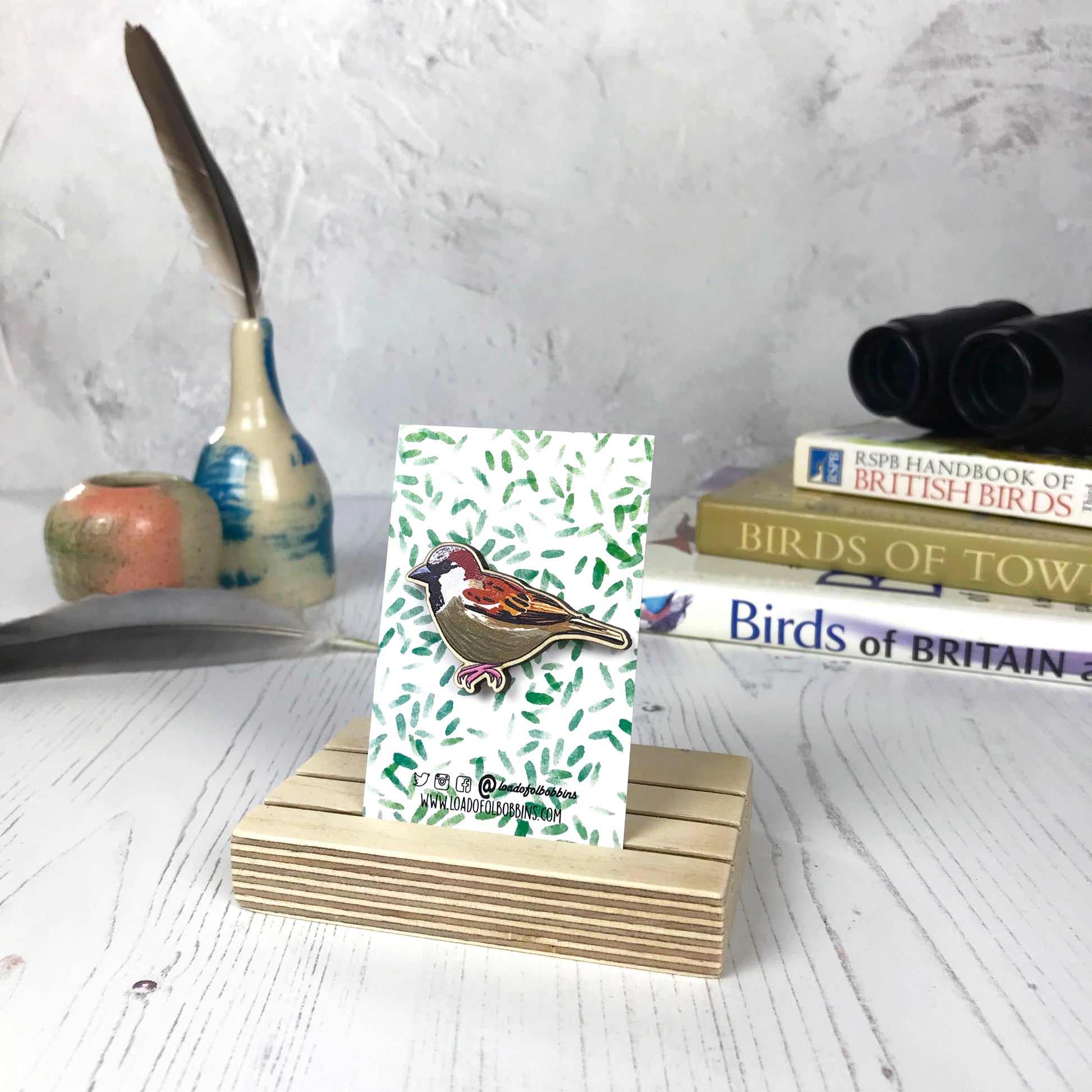 A laser cut wooden brooch showing an illustration of a male sparrow by loadofolbobbins. The brooch is mounted on a backing card with a repeat abstract watercolour leaf pattern. They are standing in a wooden stand and in the background you can see a pile of bird watching books and a pair of binoculars.