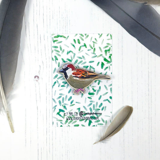 A laser cut wooden brooch showing an illustration by Loadofolbobbins of a male sparrow. The brooch is mounted on a backing card with a repeat abstract watercolour leaf pattern.