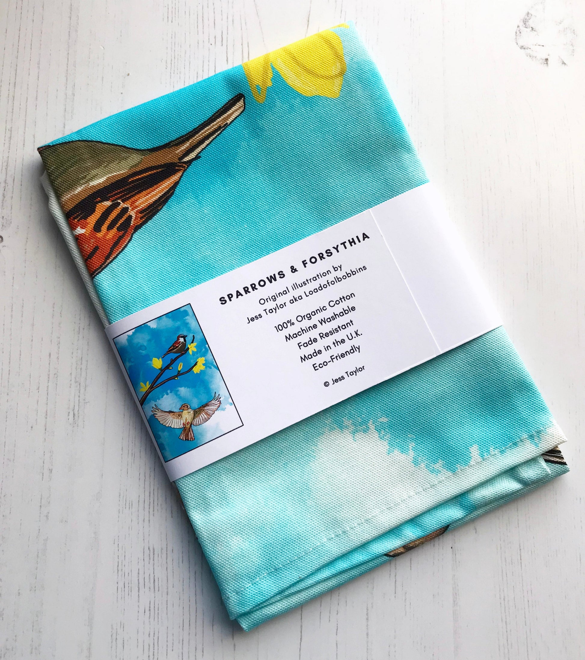 Text on belly band around a folded teatowel says. Sparrow and Forsythia tea towel. Original llustration by Jess Taylor aka Loadofolbobbins. 100% organic cotton, machine washable, fade resistant,made in the UK, Eco friendly.