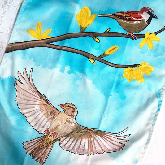 A close up of a tea towel printed with and original loadofolbobbins illustration. It shows a male sparrow perched on a branch of forsythia with bright yellow blooms. Underneath is a flying female  sparrow with wings outstretched. The background is a turquoise inky wash.