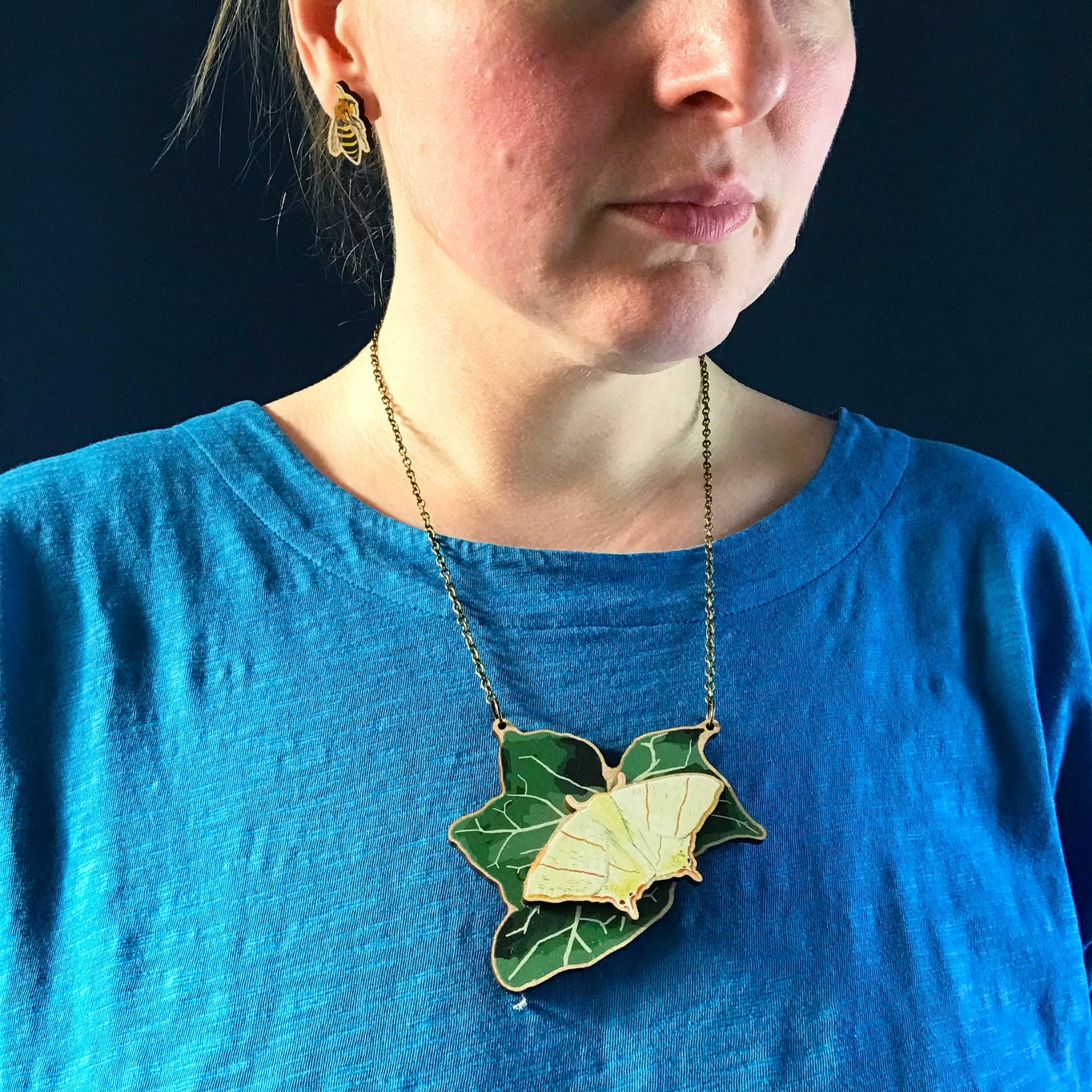 Swallowtail Moth and Ivy necklace by Loadofolbobbins. A laser cut wooden necklace that shows a large ivy leaf with a swallowtail moth on. The necklace hangs on an antique bronze chain. It is being worn by me a white woman wearing a light blue top. You can just see the bottom of my face and neck.