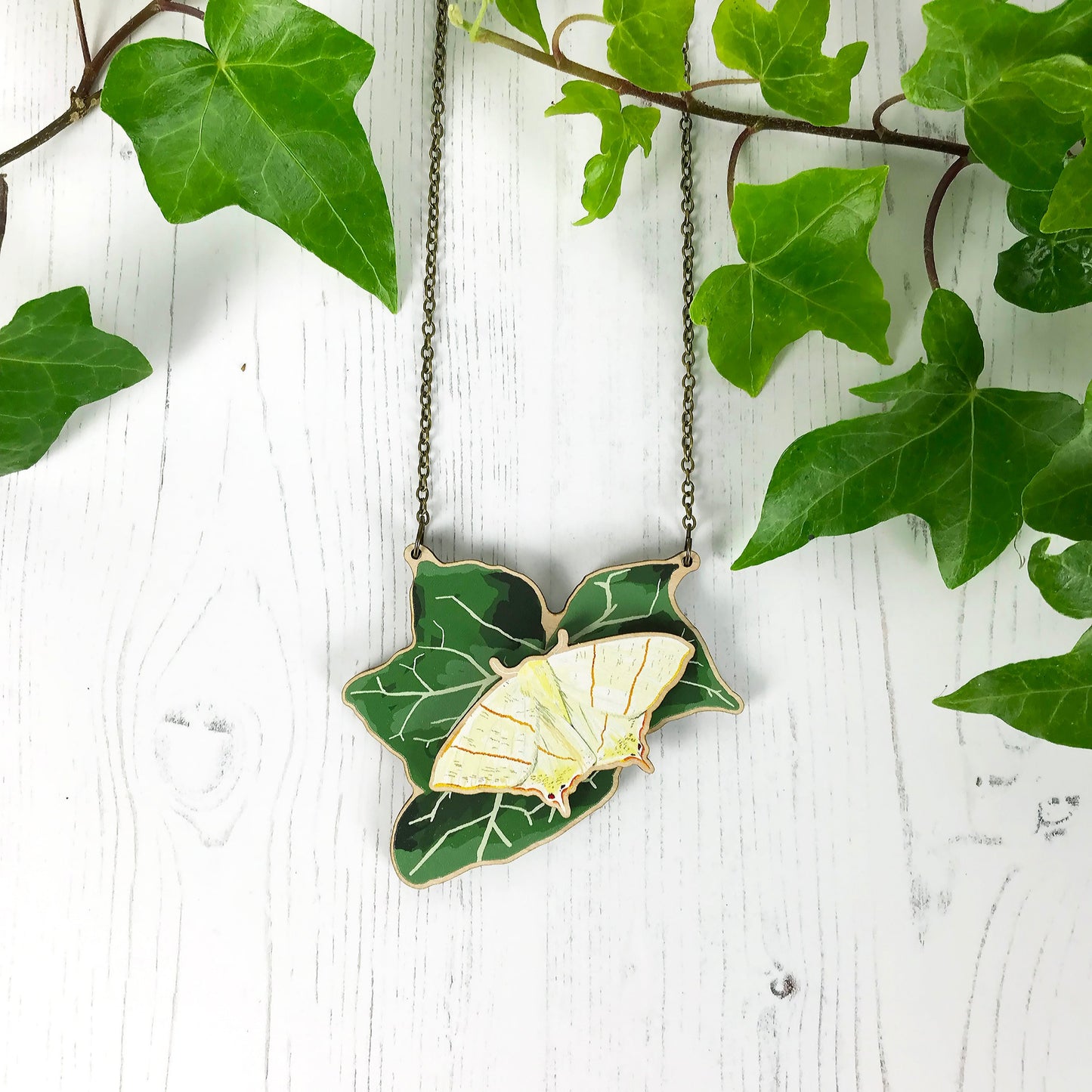 A laser cut wooden statement necklace showing an illustration of an dark green ivy leaf and Swallowtail Moth by loadofolbobbins. The moth stands proud of the necklace in a 3D relief effect.  The wooden necklace is hanging from an antique bronze cable chain.