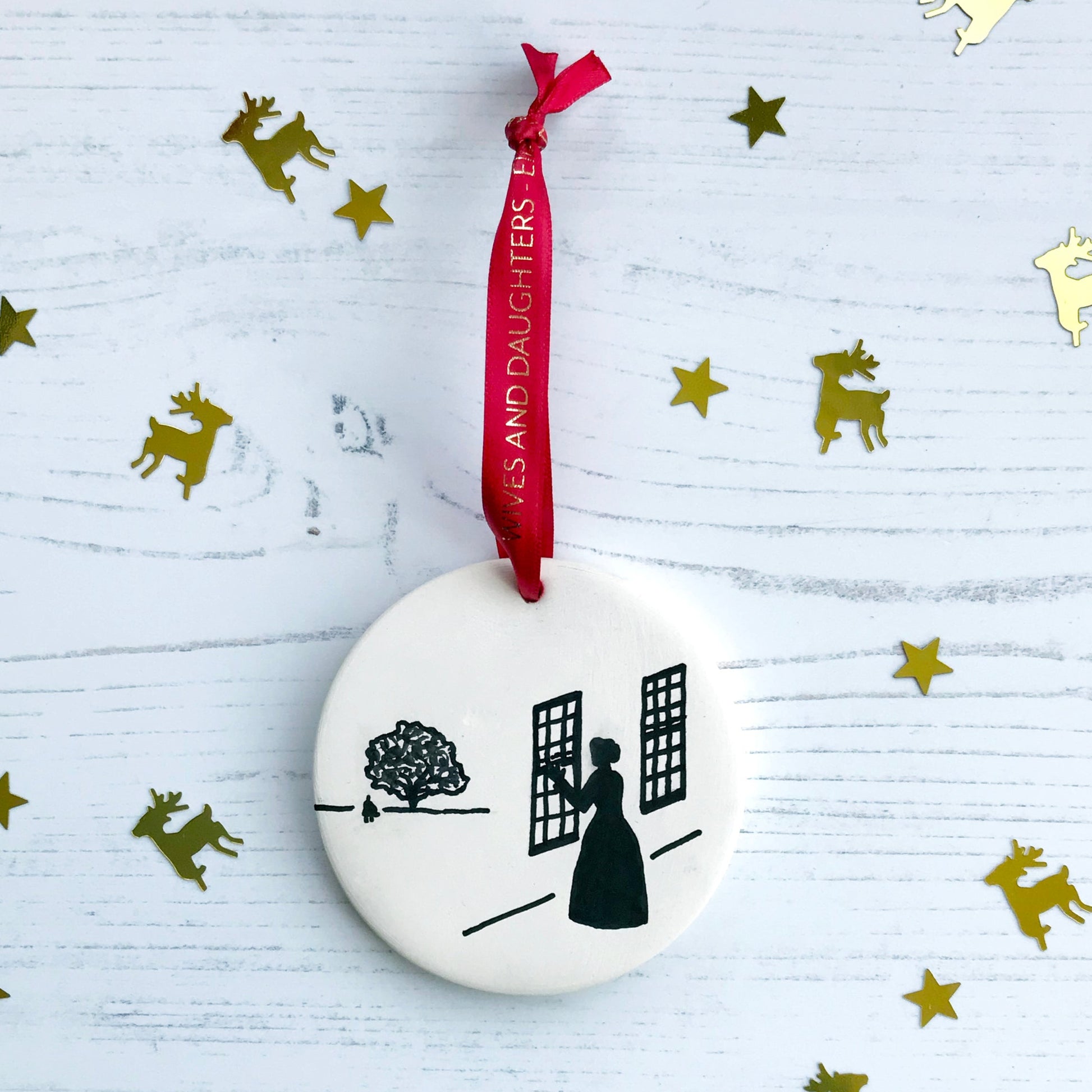 A circular white ceramic hanging decoration showing a hand painted black silhouette scene inspired the novel Wives and Daughters by Elizabeth Gaskell. The scene shows Molly looking out of the window at Roger standing under a large tree. It hangs from a satin red ribbon with gold foil lettering that says Wives and Daughters - Elizabeth Gaskell.