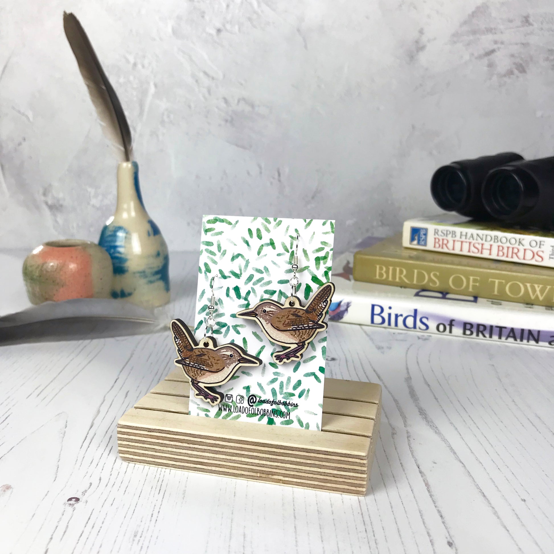 A pair of laser cut wooden dangle earrings showing an illustration of a wren bird by loadofolbobbins. The earrings are mounted on a backing card with a repeat abstract watercolour leaf pattern. They are standing in a wooden stand and in the background you can see a pile of bird watching books and a pair of binoculars.