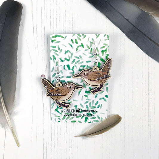 A pair of laser cut wooden dangle earrings showing an illustration by Loadofolbobbins of a wren. The earrings are mounted on a backing card with a repeat abstract watercolour leaf pattern.