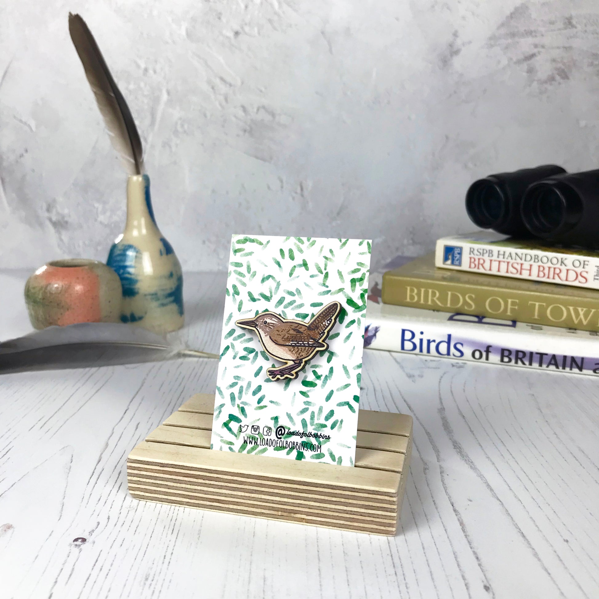 A laser cut wooden brooch showing an illustration of a wren bird by loadofolbobbins. The brooch is mounted on a backing card with a repeat abstract watercolour leaf pattern. They are standing in a wooden stand and in the background you can see a pile of bird watching books and a pair of binoculars.