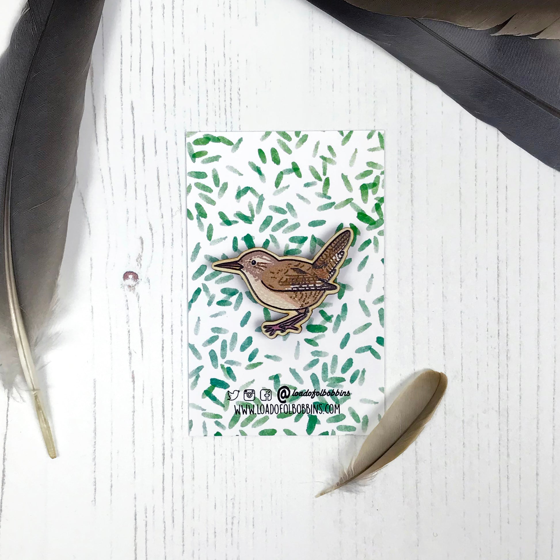 A laser cut wooden brooch showing an illustration by Loadofolbobbins of a wren. The brooch is mounted on a backing card with a repeat abstract watercolour leaf pattern.