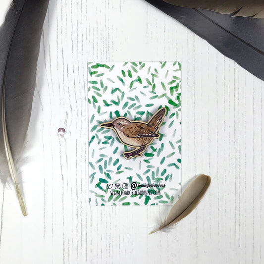 A laser cut wooden brooch showing an illustration by Loadofolbobbins of a wren. The brooch is mounted on a backing card with a repeat abstract watercolour leaf pattern.