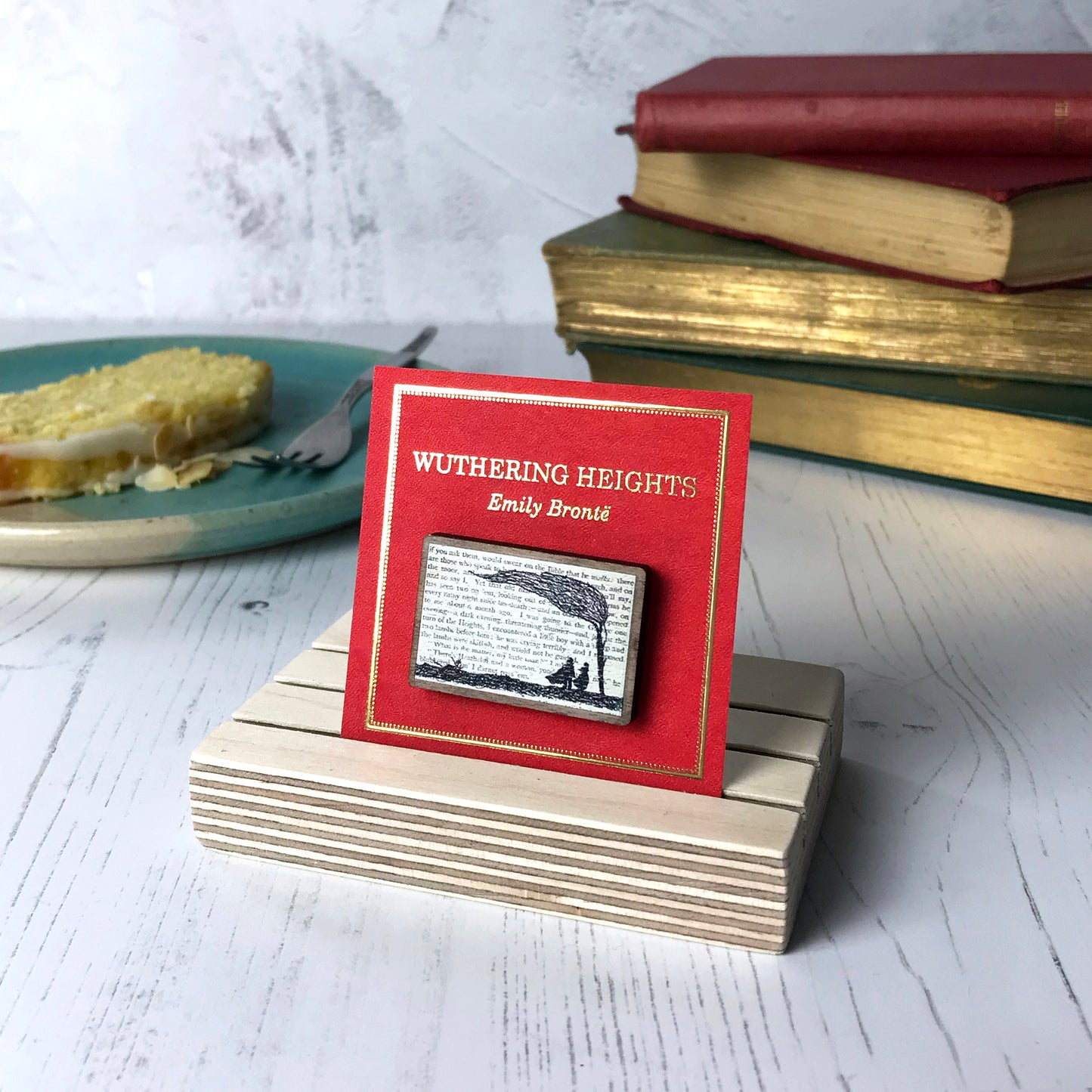 A laser cut wooden brooch by loadofolbobbins showing a silhouette scene inspired by the novel Wuthering Heights by Emily Brontë. The scene shows a silhouette scene of a man and woman under a windswept tree, Heathcliffe and Cathy. The scene is printed against original text from the novel. The necklace is mounted on a red backing card with gold foil lettering detail and made to look like the front of a book.