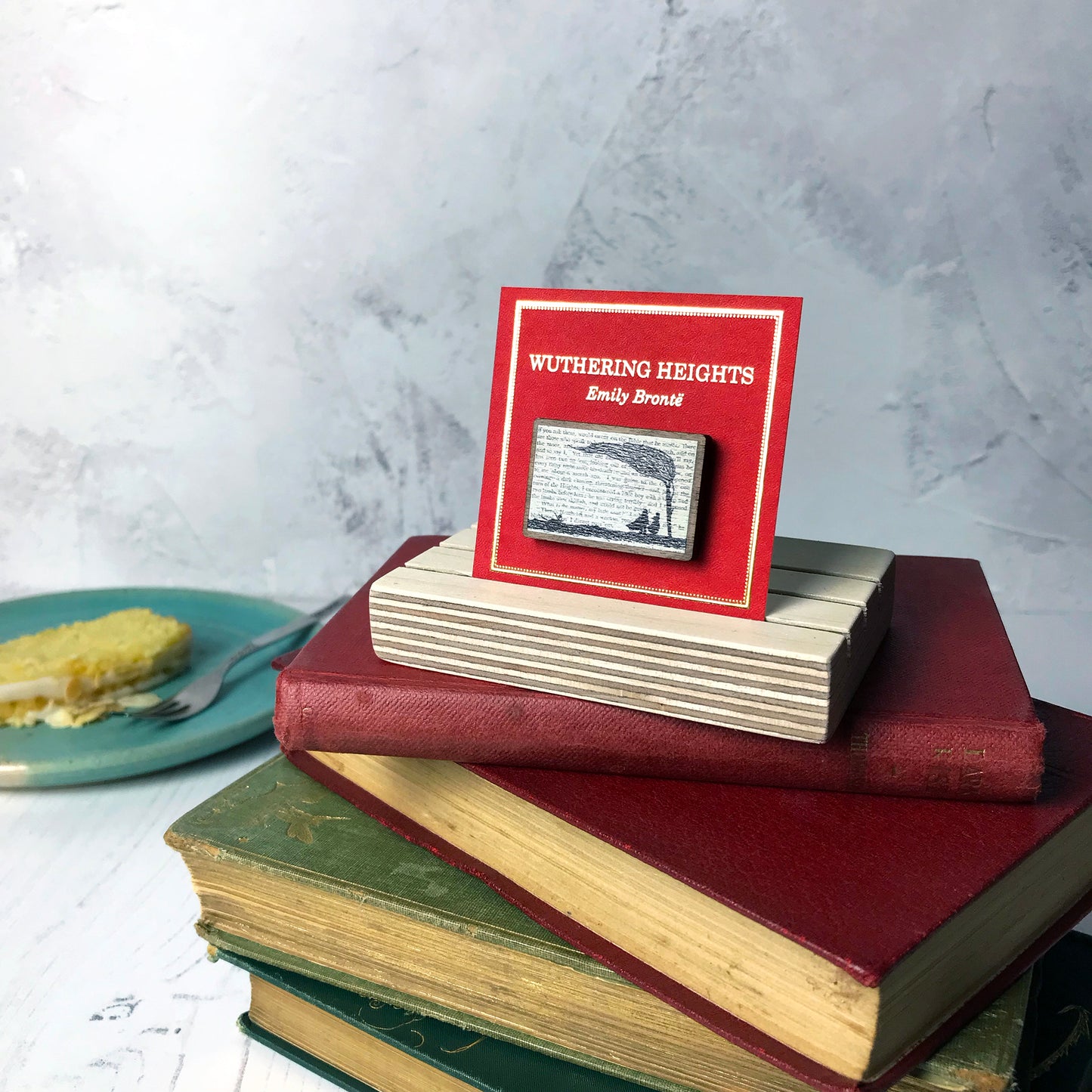 A laser cut wooden brooch by loadofolbobbins showing a silhouette scene inspired by the novel Wuthering Heights by Emily Brontë. The scene shows a silhouette scene of a man and woman under a windswept tree, Heathcliffe and Cathy. The scene is printed against original text from the novel. The necklace is mounted on a red backing card with gold foil lettering detail and made to look like the front of a book.