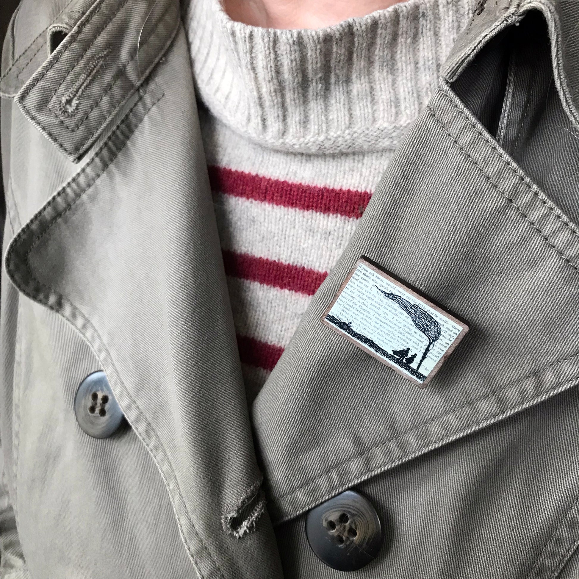 A laser cut wooden brooch by loadofolbobbins showing a silhouette scene inspired by the novel Wuthering Heights by Emily Brontë. The scene shows a silhouette scene of a man and woman under a windswept tree, Heathcliffe and Cathy. The scene is printed against original text from the novel. The brooch is being worn on the lapel of a jacket.