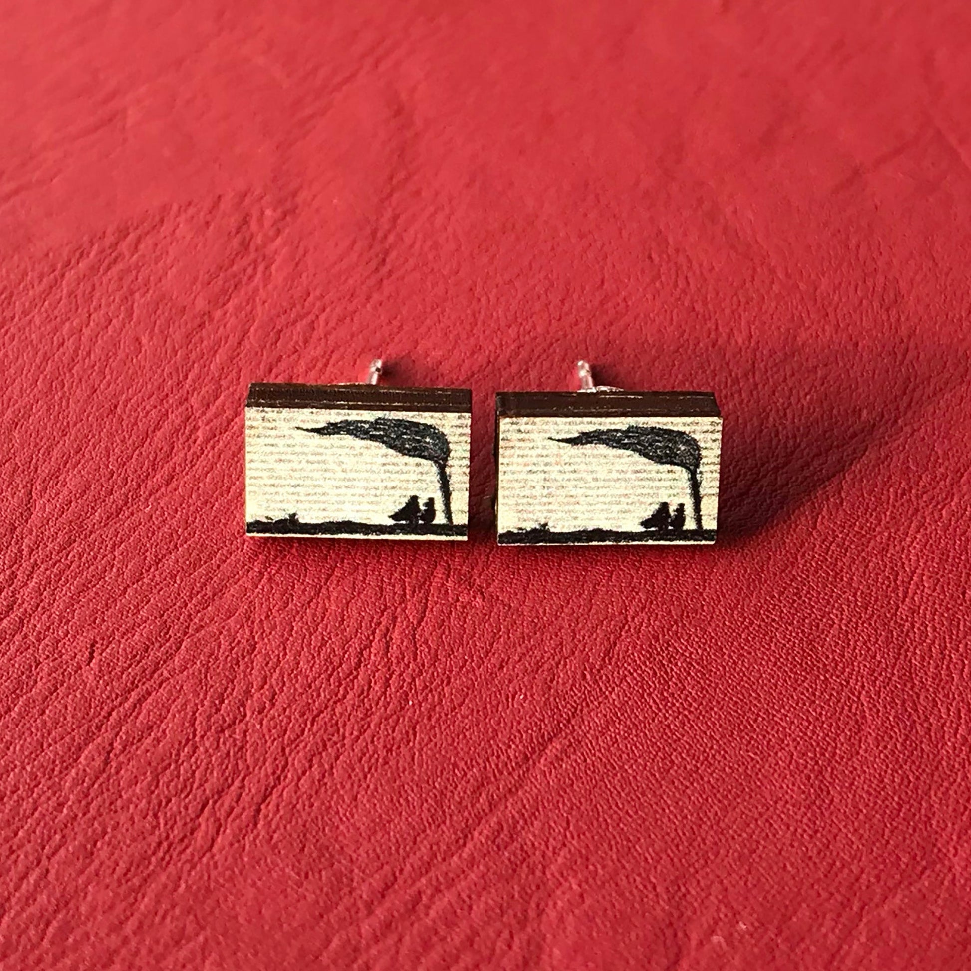 A laser cut wooden stud earrings by loadofolbobbins showing a silhouette scene inspired by the novel Wuthering Heights by Emily Brontë. The scene shows a silhouette scene of a man and woman under a windswept tree, Heathcliffe and Cathy. The scene is printed against original text from the novel. 