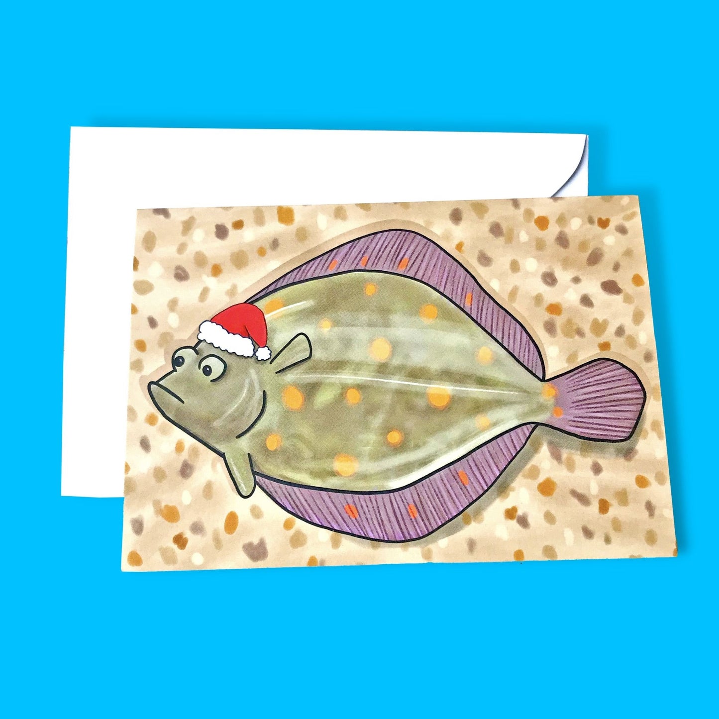 An A6 greetings card showing a slightly comic illustration by Loadofolbobbins of a Plaice fish lying on a sandy pebbly beach wearing a Santa hat. The plaice has a slightly grumpy expression. It is a greeny brown in colour with orange spot markings with a light watery sheen on its surface. It is a kind of rounded diamond shape and has both eyes on the same side of its head.
