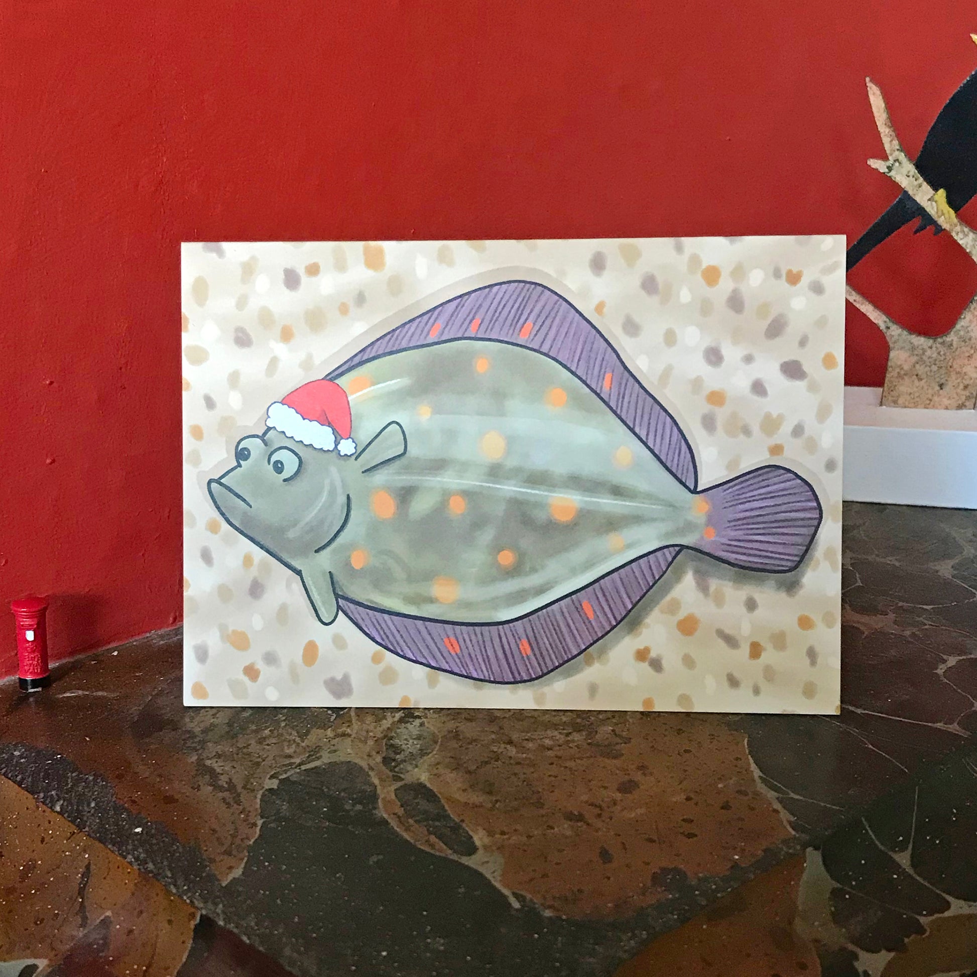 An A6 greetings card showing a slightly comic illustration by Loadofolbobbins of a Plaice fish lying on a sandy pebbly beach wearing a Santa hat. The plaice has a slightly grumpy expression. It is a greeny brown in colour with orange spot markings with a light watery sheen on its surface. It is a kind of rounded diamond shape and has both eyes on the same side of its head. The card is displayed on a mantelpiece.