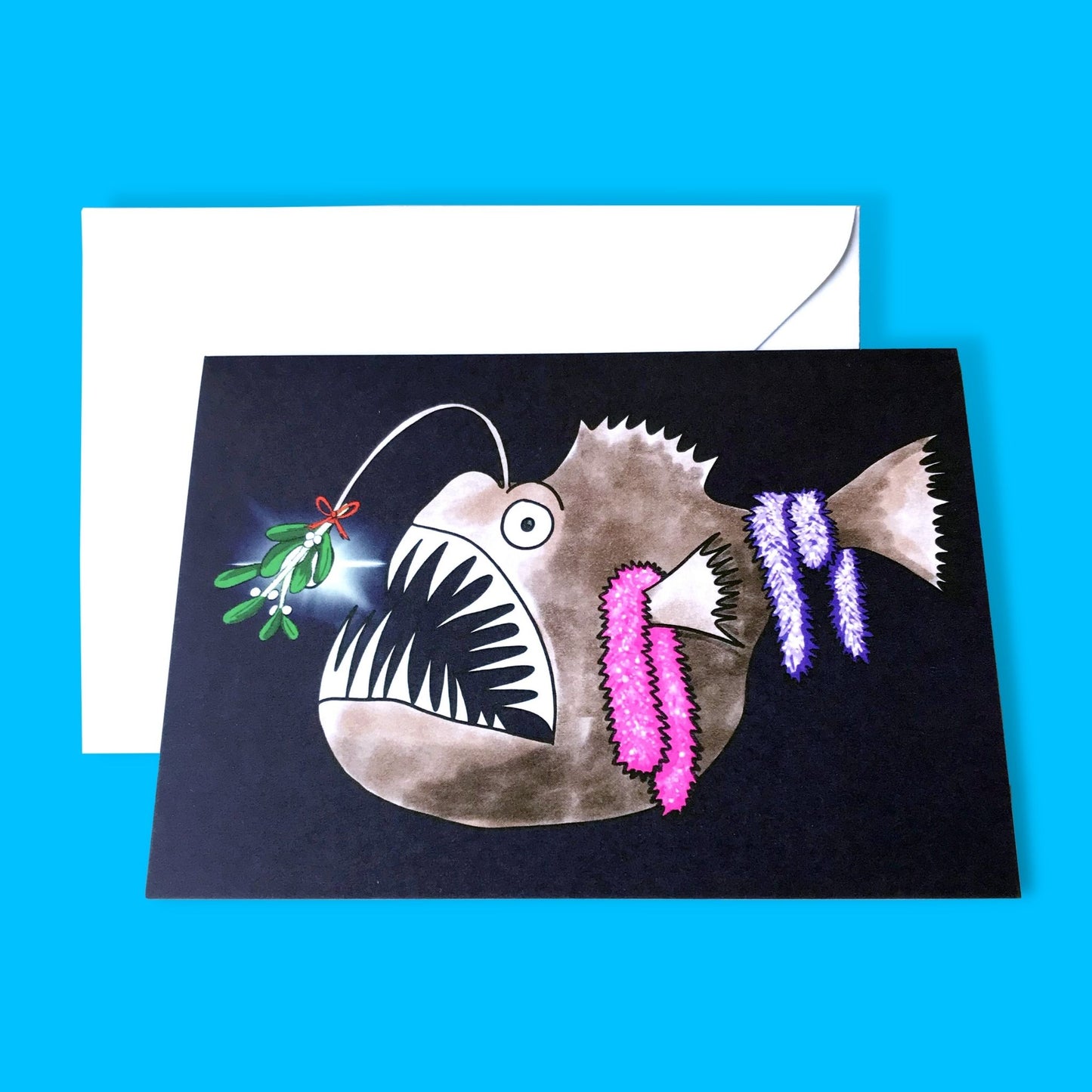 An A6 greetings card showing an illustration by Loadofolbobbins of an Anglerfish in the dark depths of the ocean. It is done in a cartoonish style. There’s a sprig of mistletoe attached to the bioluminescent lure that hangs from its head and some sparkly pink and purple tinsel hanging from its fins and tail. Mainly brown in colour it has a large gaping mouth full of very sharp pale teeth and a big round eye. 