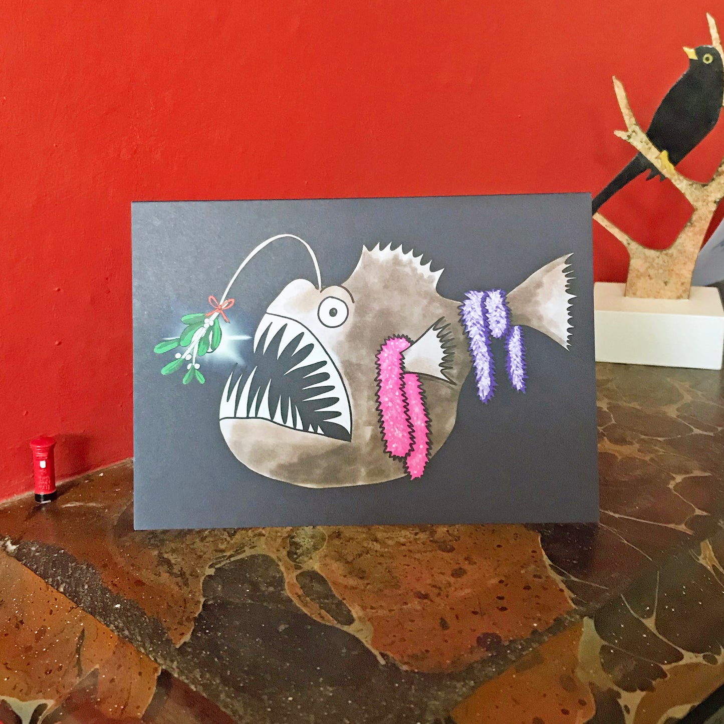 An A6 greetings card showing an illustration by Loadofolbobbins of an Anglerfish in the dark depths of the ocean. It is done in a cartoonish style. There’s a sprig of mistletoe attached to the bioluminescent lure that hangs from its head and some sparkly pink and purple tinsel hanging from its fins and tail. Mainly brown in colour it has a large gaping mouth full of very sharp pale teeth and a big round eye. The card is displayed on a mantelpiece.