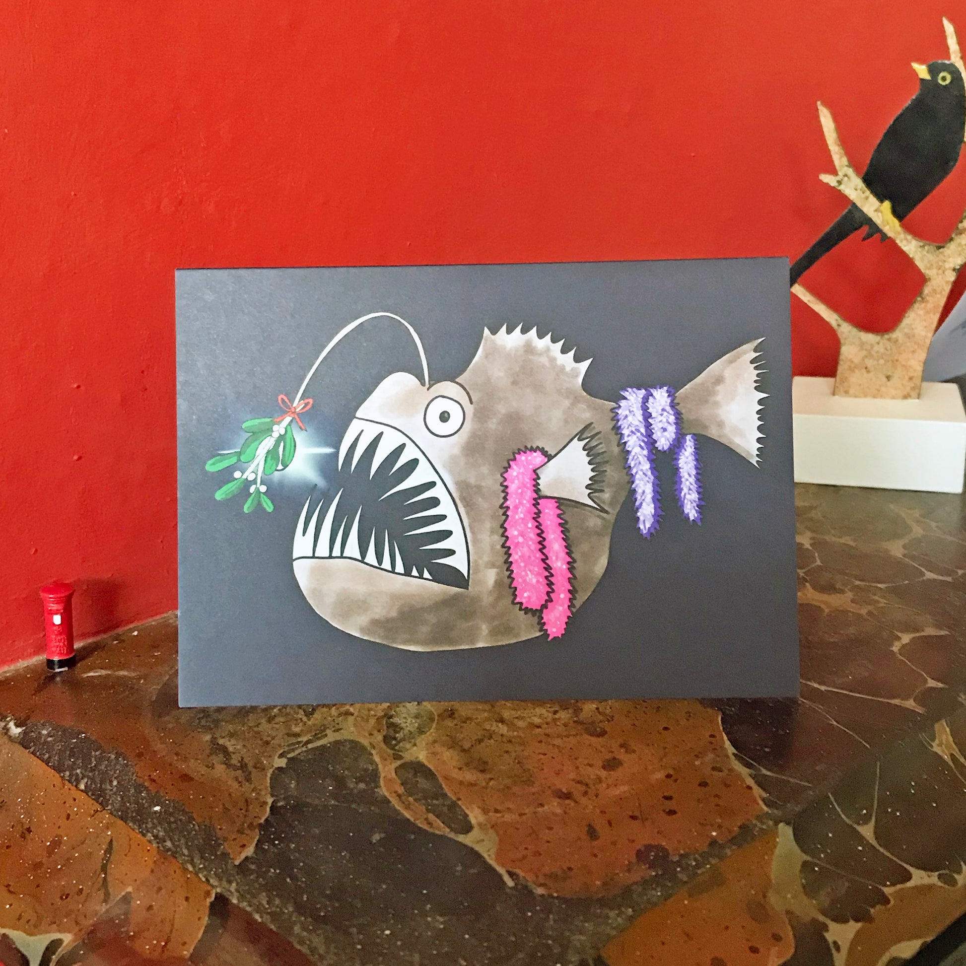 An A6 greetings card showing an illustration by Loadofolbobbins of an Anglerfish in the dark depths of the ocean. It is done in a cartoonish style. There’s a sprig of mistletoe attached to the bioluminescent lure that hangs from its head and some sparkly pink and purple tinsel hanging from its fins and tail. Mainly brown in colour it has a large gaping mouth full of very sharp pale teeth and a big round eye. The card is displayed on a mantelpiece.