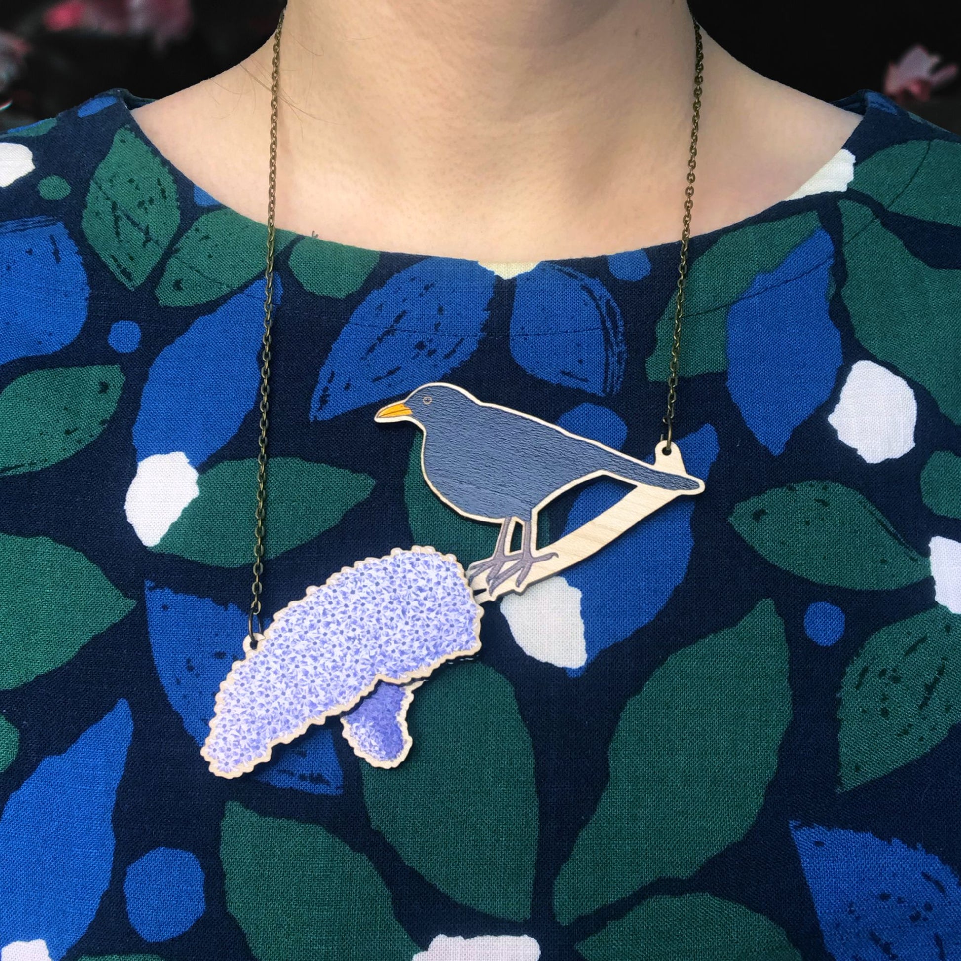 A laser cut wooden charm necklace showing an illustration of a male blackbird perching on a branch with lilac blossoms by loadofolbobbins. The lilac blossom flowers stand proud of the necklace in a 3D relief effect.  The wooden necklace is hanging from an antique bronze cable chain. It is being worn by me a white woman wearing a blue and green leaf patterned top. You can just see the bottom of my neck.